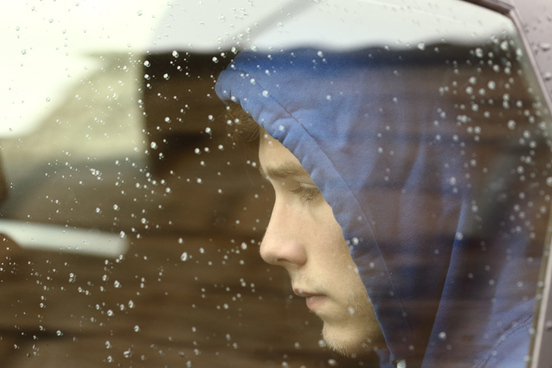 Porträtt på en nedstämd tonåring med huvan uppdragen, som ser ut genom ett regnigt bilfönster.