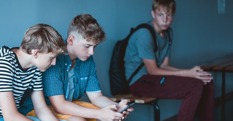 Tre unga killar sitter på skolbänkar intill väggen. Två tittar ner i en telefon och den tredje sitter och tittar på den andra killarna.
