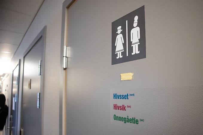 En toalettdörr med en skylt med två samiska figurer på, en med kvinnligt kodade kläder och en med manligt kodade kläder. Under står toalett på nordsamiska, lulesamiska och sydsamiska.