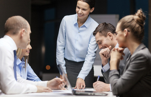 En leende chef i blå skjorta står i änden av ett konferensbord och pekar på ett papper för fyra glada kollegor.