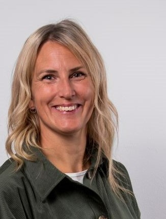 Porträttbild på en person med ljust långt hår, grön skjorta och en vit t-shirt som skymtar i halsringningen. Personer ler och tittar in i kameran.