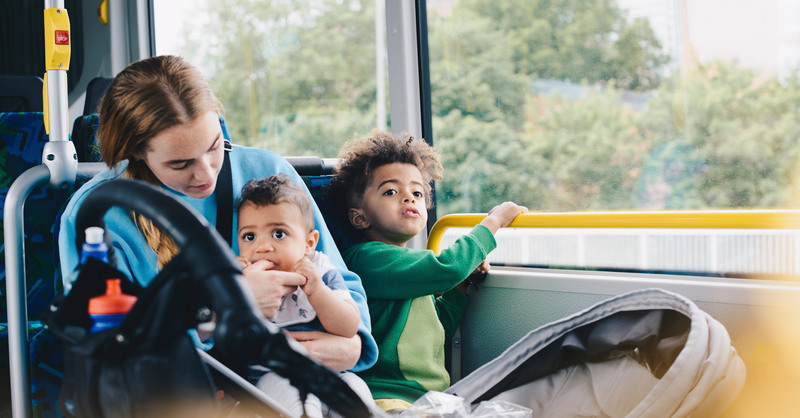 En vuxen sitter med två små barn på en buss med en barnvagn framför sig.