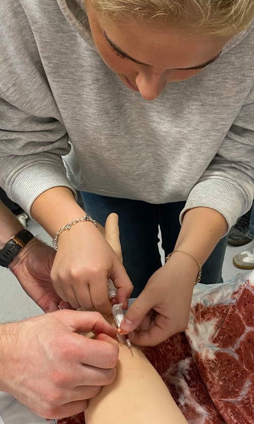 En gymnasieelev övar på att sätta en nål i en arm-attrapp under ett studiebesök på Kliniskt träningscentrum, Region Jämtland Härjedalen. En utbildningsledare står bredvid och instruerar.