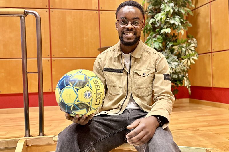 Leende mörkhårig person i glasögon sitter i en trappa med en gulblå fotboll i höger hand