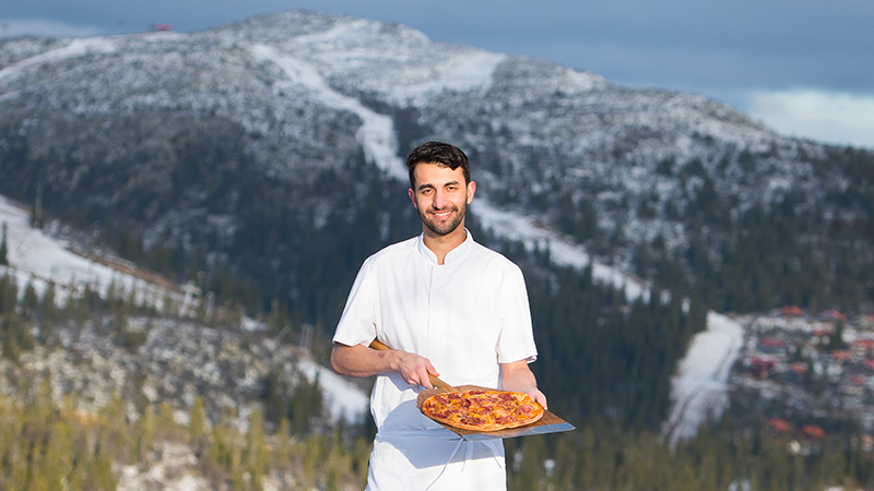 Porträttbild av en pizzabagare i vita bagarkläder som visar upp en pizza på sin stekspade, ståendes på fjället med ett halvsnöigt fjäll i bakgrunden.