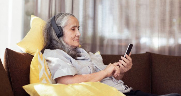 Kvinna sitter i soffa med hörlurar på och tittar på sin mobiltelefon. 