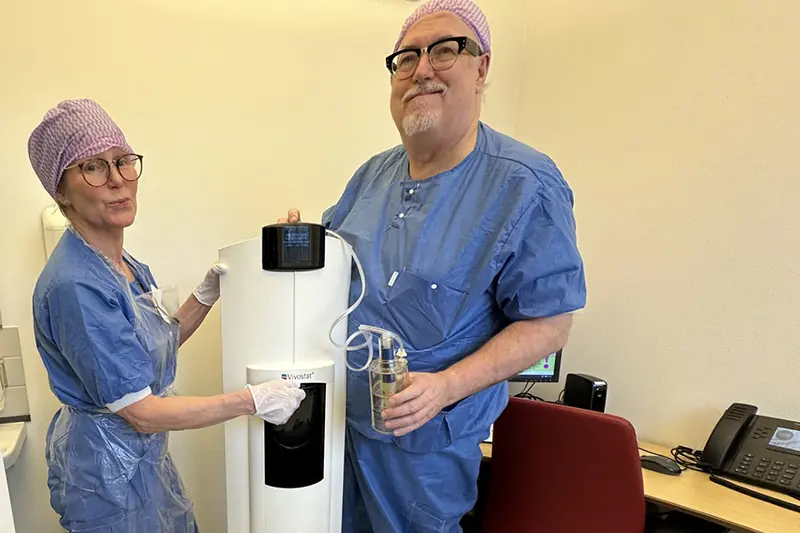 Två personer i blå operationskläder och hättor på sina huvuden står vid en medicinteknisk apparat.