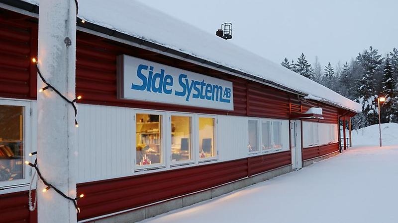 Side Systems röda plåtbyggnad med en stor skylt med företagsnamnet och en julig ljusslinga runt flaggstången i förgrunden.