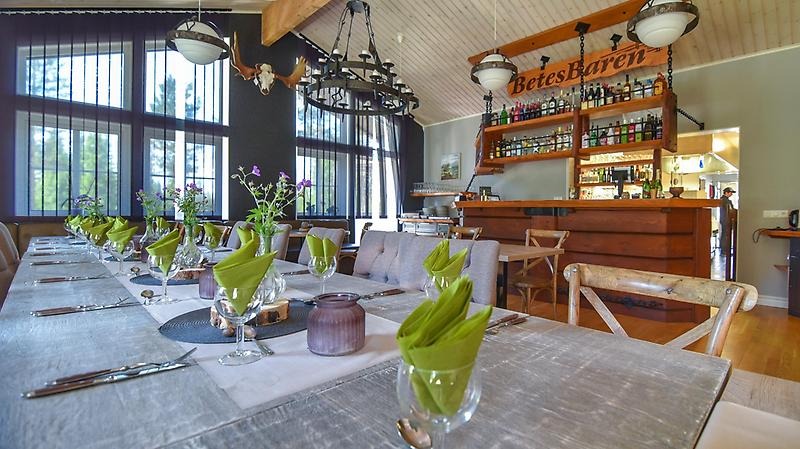 Restaurangbord uppdukat med vinglas och finporslin, framför en bar med skylten "Betesbaren" och stora fönster med ljusinsläpp.