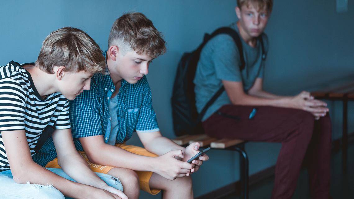 Två tonårspojkar som sitter på skolbänk och tittar i en mobiltelefonen. Ledsen ensam pojke med ryggsäck som sitter på samma bänk en bit ifrån dem.
