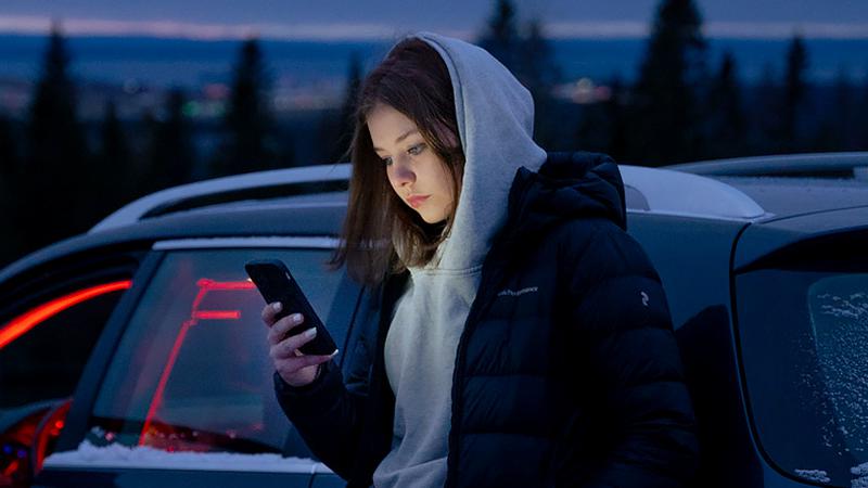 Ung person i luvtröja står lutad mot en A-traktor i kvällsmörker och tittar på sin mobiltelefon.