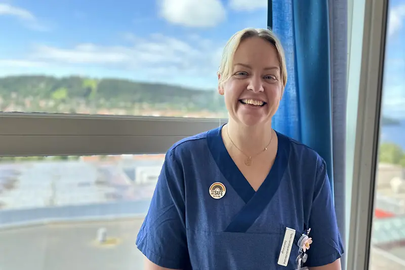 En person i ljust hår uppsatt i nacken och blå kläder står vid ett fönster med utsikt över Frösön. Hen ler in i kameran.