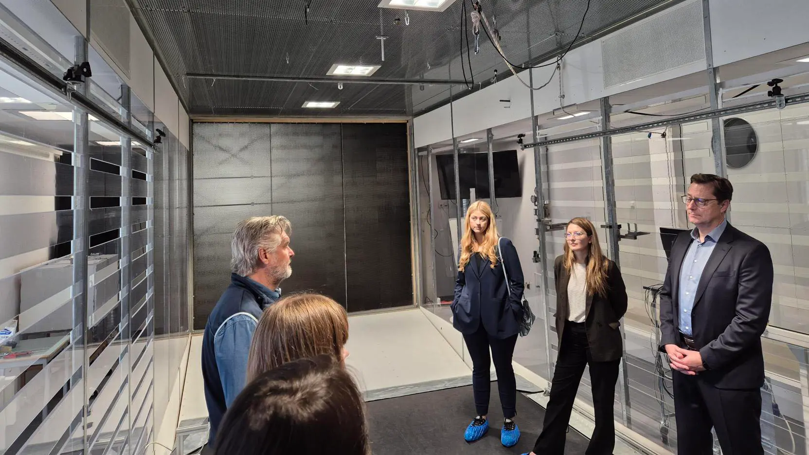 Sex personer står i en lokal som innehåller en så kallad testbädd. Längs väggarna syns hyllor, sladdar och tv-skärmar. En person ser ut att berätta om innehållet i rummet för de andra personerna som lyssnar. 