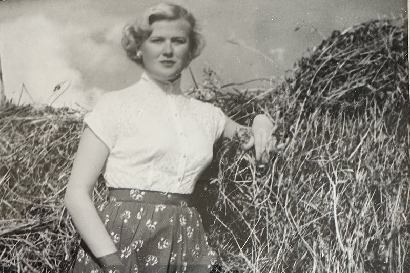 Ett svartvitt foto från tidigt 1950-tal där en blond kvinna står lutad mot en höhässja iklädd finblus, smycken och plisserad kjol.