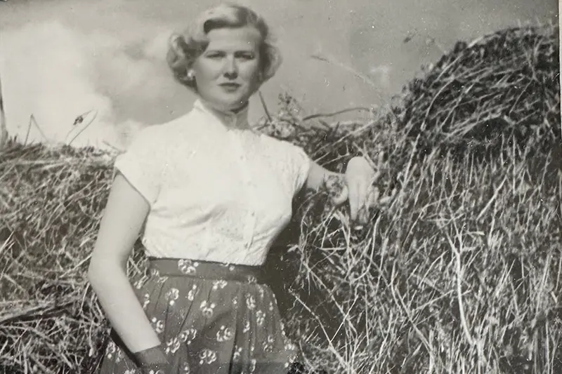 Ett svartvitt foto från tidigt 1950-tal där en blond kvinna står lutad mot en höhässja iklädd finblus, smycken och plisserad kjol.