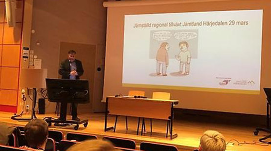 Tre åhörare lyssnar på en man som håller ett föredrag och visar en bild på en projektorduk