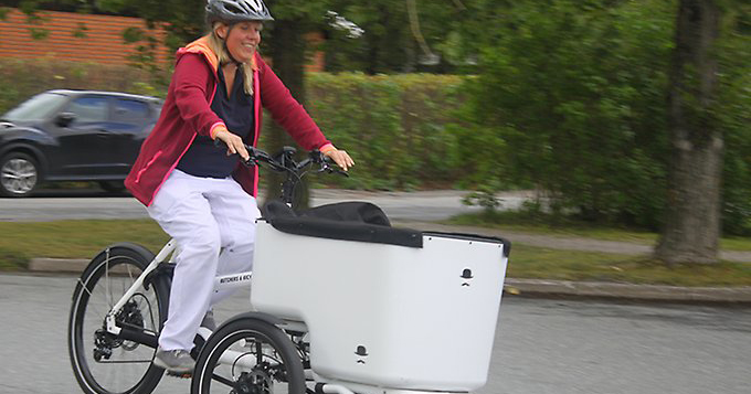 En sjuksköterska testcyklar en elcykel med packlåda framtill. Hon har cykelhjälm på huvudet.