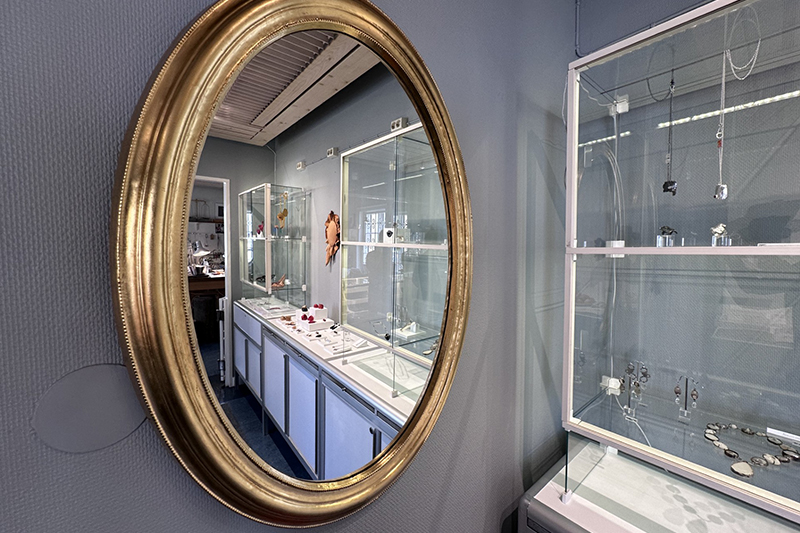 En oval spegel i guldram i vars glas man ser delar av Galleri Smedjans konsthantverk