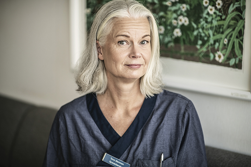 Person i blå vårdkläder och axellångt ljust hår tittar in i kameran