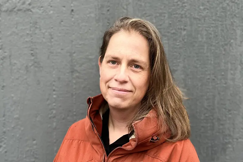 Närbild på person som ler med stängd mun. Axellångt brunt hår och rostfärgad vindjacka.