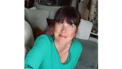 Mjukt leende person med brunt hår uppsatt i tofs och brun lugg, samt turkos tröja. I bakgrunden syns en vardagsrumsmiljö med grå soffor.