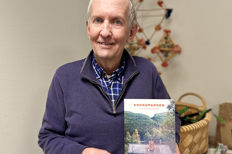 Person i blå tröja och grått hår ler mot kameran. I händerna håller hen upp en bok