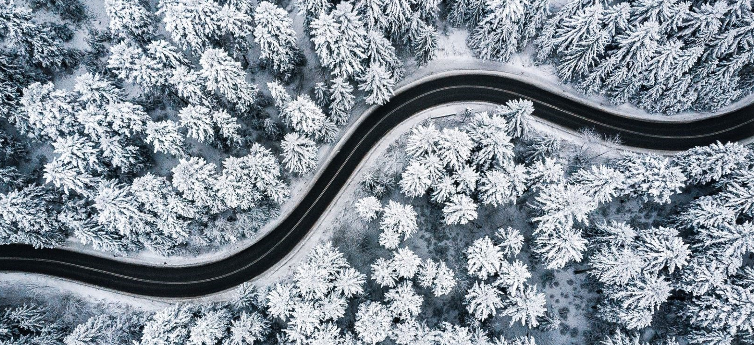 En svart asfaltsväg fotad uppifrån, slingrandes genom ett landskap av snöiga granar.