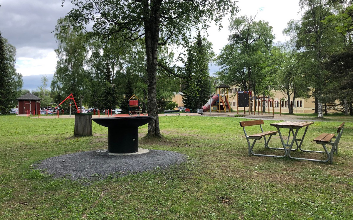 Bild på en utegrill med en bänk bredvid. En lekplats i bakgrunden.