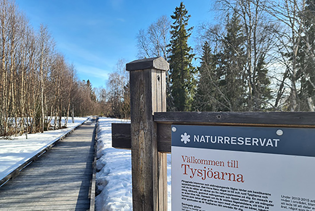 Foto på en träspång ut i naturen. En skylt till höger som det står Välkommen till Tysjöarna på.