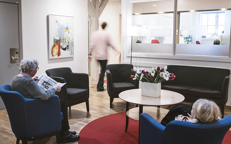 Två personer, varav en är äldre och en är ett barn, sitter varsin fåtölj i ett väntrum. Väntrummet är möblerat med soffa och flera fåtöljer samt ett soffbord med blommor på. Den äldre person läser tidningen. Någon passerar förbi i bakgrunden och på väggen hänger ett konstverk.