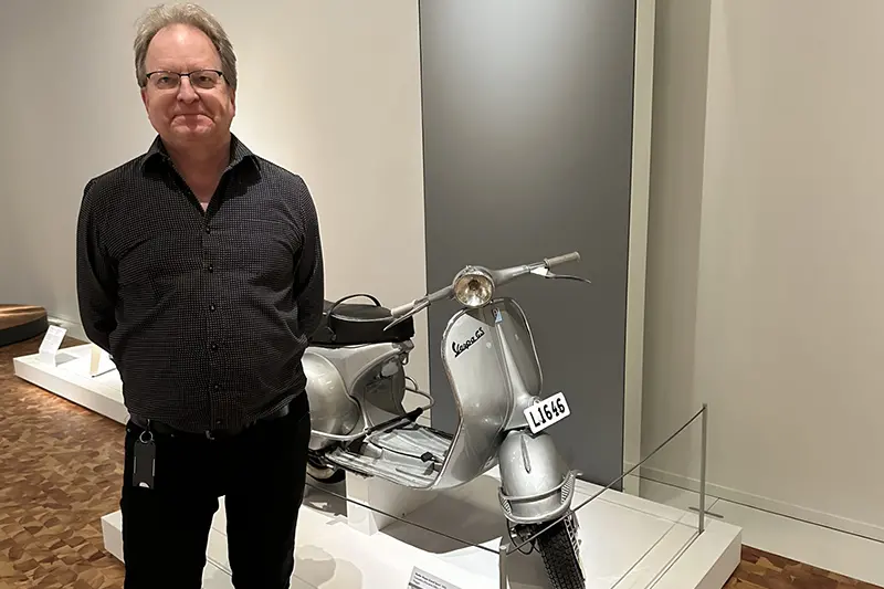 En person i jeans och skjorta och kort hår står framför en vespa från 50-talet