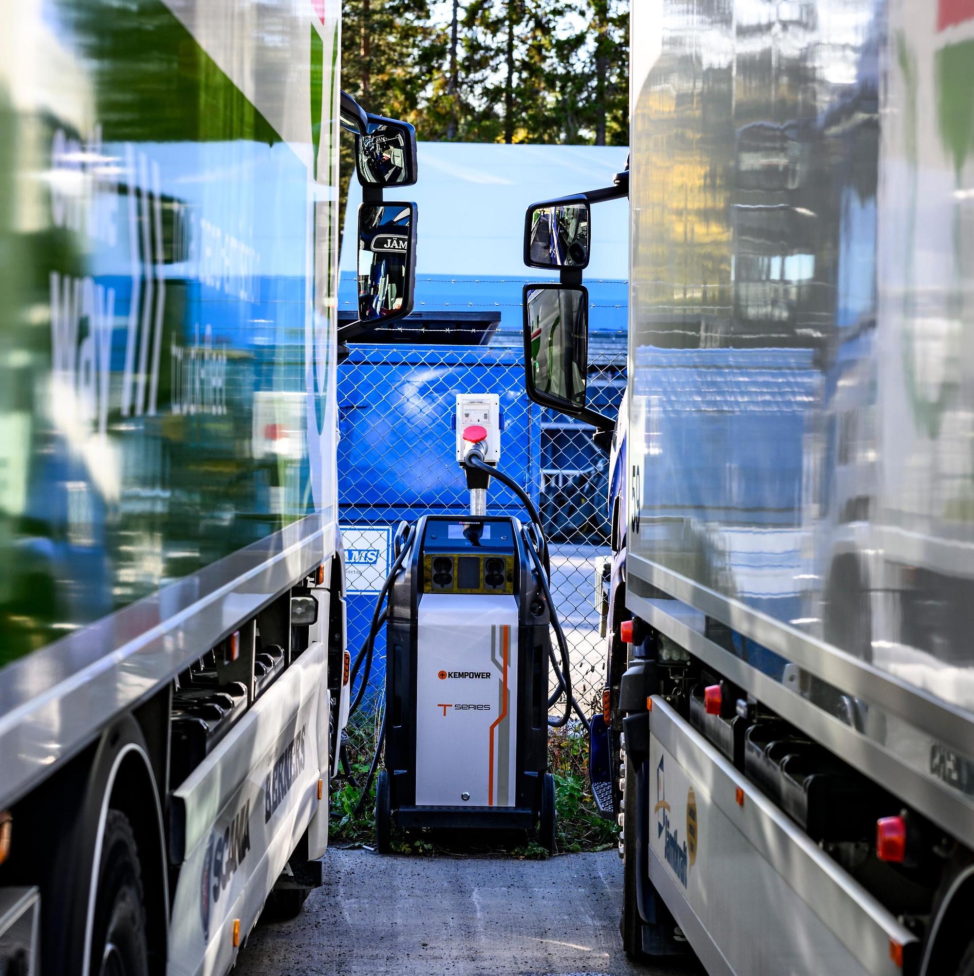 Närbild på ett ladduttag med en sladd inkopplad, mellan två parkerade eldrivna lastbilar.