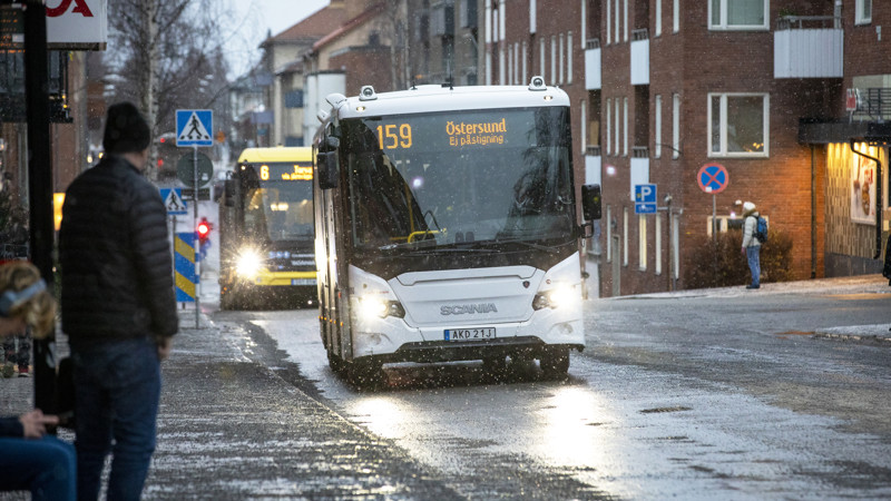 En buss med texten "159 Östersund, ej påstigning" på skylten. Den kör längs en regnig gata i centrala Östersund, betraktad av personer i förgrunden vid en busskur.