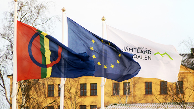 Flaggor vajandes utanför Regionens hus i Östersund: samiska flaggan, EU-flaggan och en flagga med Region Jämtland Härjedalens logga på.