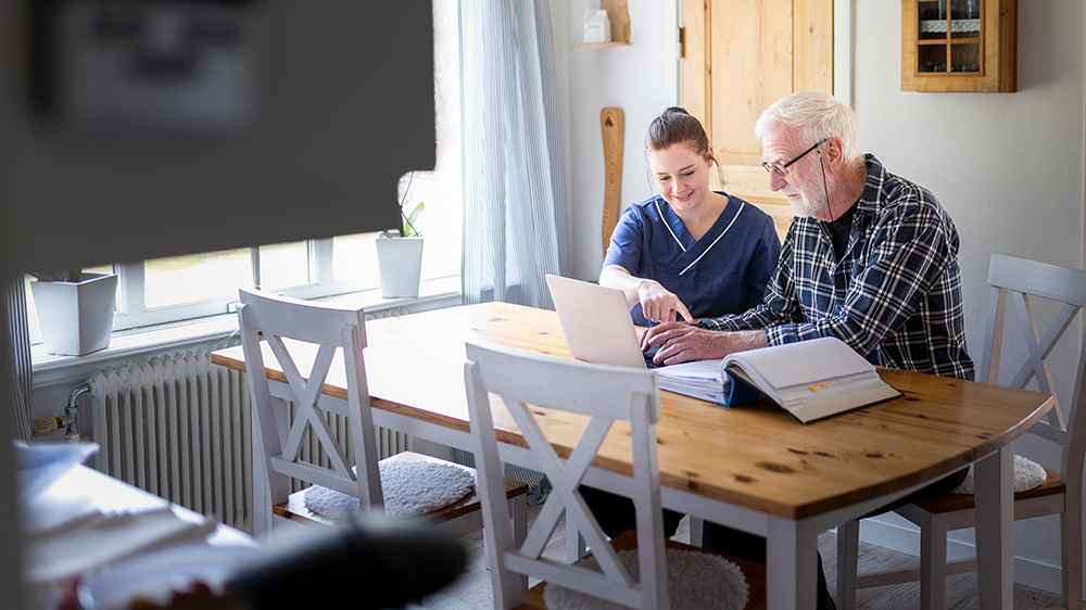 Hemtjänstpersonal hjälper en person med datorn hemma vid dennes köksbord.
