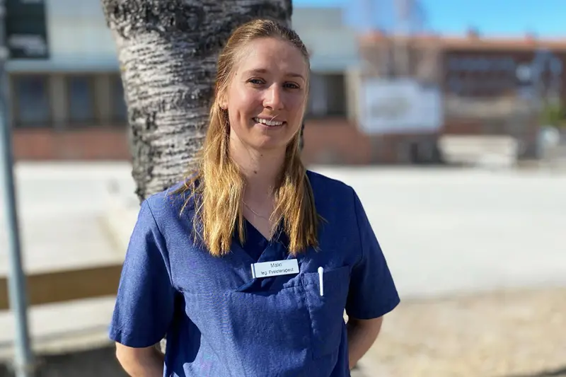 En person står lutad mot en björk i solskenet. klädd i blå arbetskläder