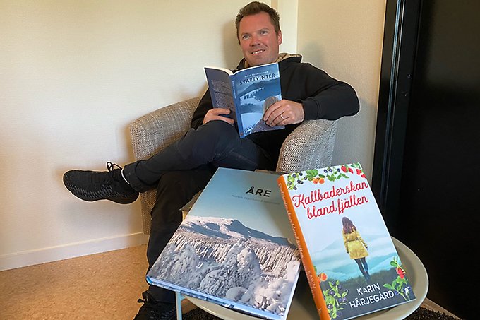 En person sitter i en fåtölj med en öppen bok i händerna. Personen tittar in i kameran och ler.