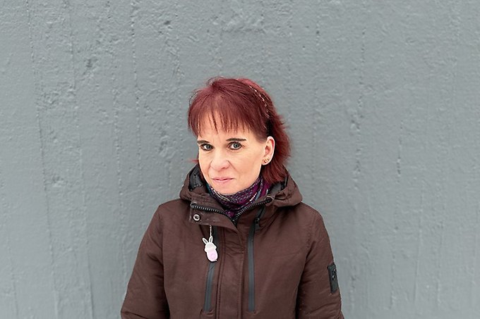 Person i vinterjacka och rött hår står mot en grå stenvägg och tittar in i kameran