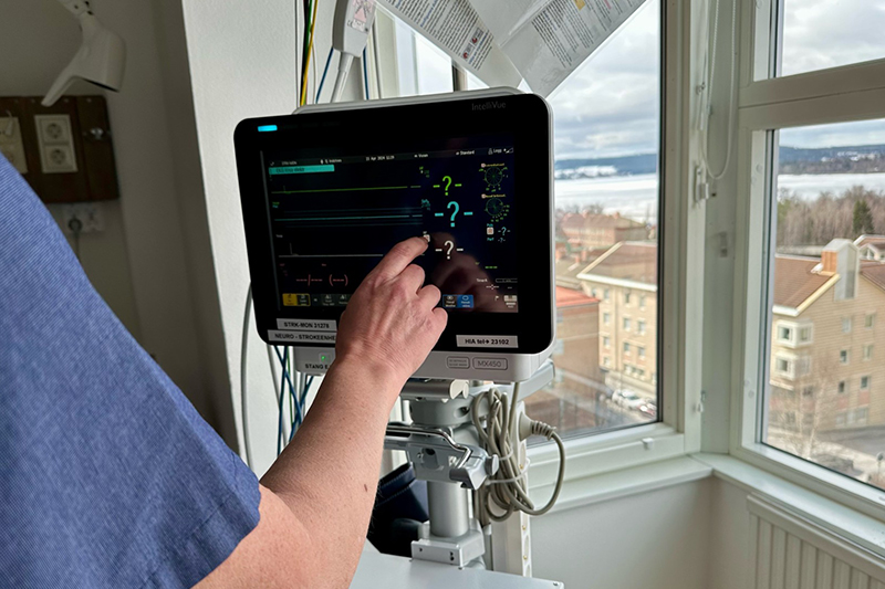 En arm kommer upp i neder vänsterkant av bilden och trycker på en skärm med patientdata. Skärmen står invid ett fönster med utsikt från höghuset på sjukhuset.