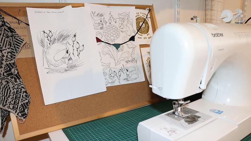 Symaskin och en anslagstavla bakom med olika skisser och stickmönster.