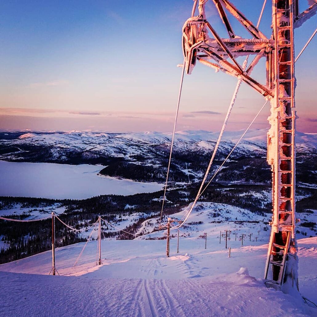 Skymningsljus över Stora Blåsjöns skidlift, med liftspår och liftstolpe i förgrunden samt milsvid utsikt över vinterfjäll i bakgrunden.