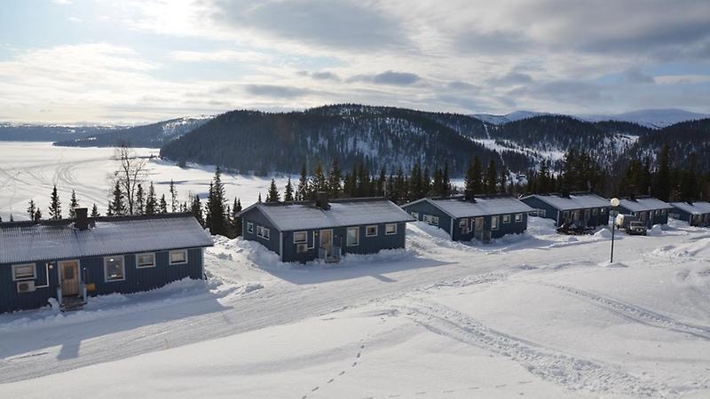 Blå campingstugor i Stora Blåsjön, med snöig omgivning och med sjön och skogsbeklädda fjäll i bakgrunden.