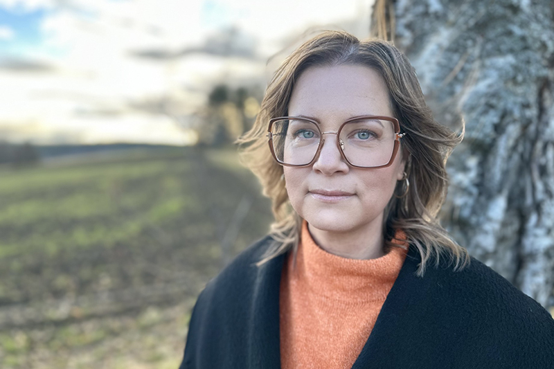 Bild på en person i axellångt brunt hår och glasögon som står lutad mot en björkstam och tittar in i kameran