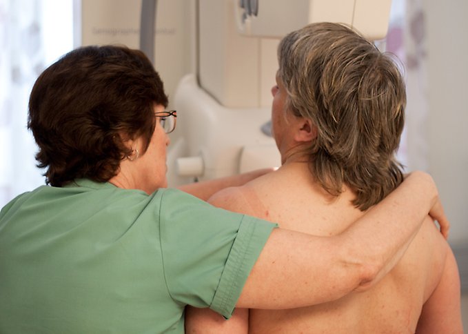 Två kvinnor står med ryggarna mot kamera. Den ena kvinnan gör en mammografiundersökning på den andra kvinnan. 