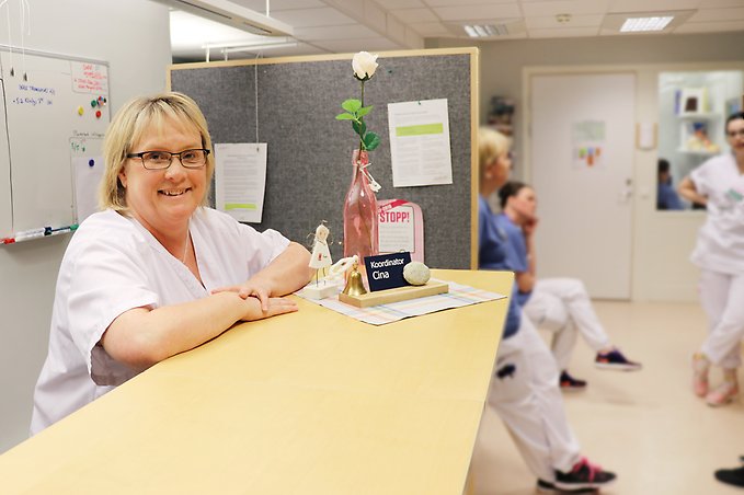 en kvinna står med armarna vilande på en disk, i bakgrunden pratar vårdpersonal med varandra