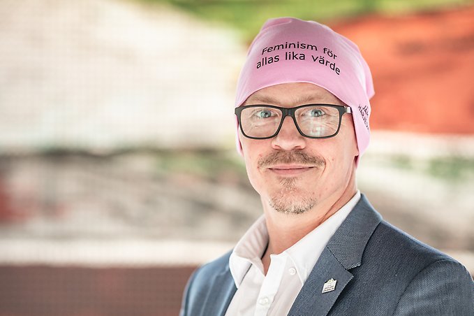 porträtt på en man i glasögon, han har en rosa mössa på huvudet på vilken det står "feminism för allas lika värde"
