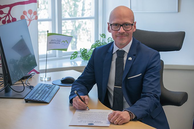 Regionfullmäktiges ordförande Thomas Andersson sitter vid ett skrivbord och skriver under ett papper. 