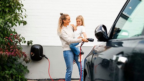 En förälder kopplar in laddsladden i sin elbil, hållandes sitt leende barn i famnen. Bilden symboliserar en elektrifierad framtid.
