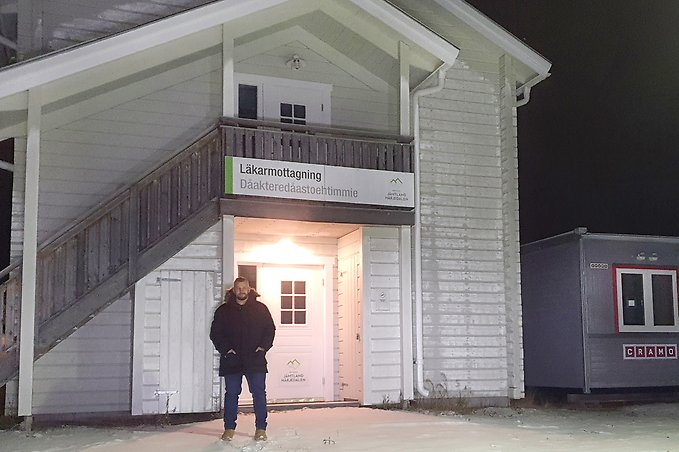en person klädd i svart vinterjacka står utanför en vitmålad träbyggnad med skylten Läkarmottagning på fasaden