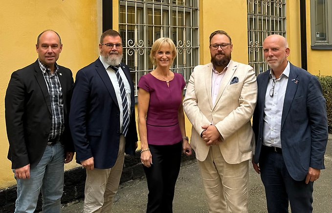 Fem personer står på rad framför en gul husvägg. Personerna ler och tittar in i kameran.